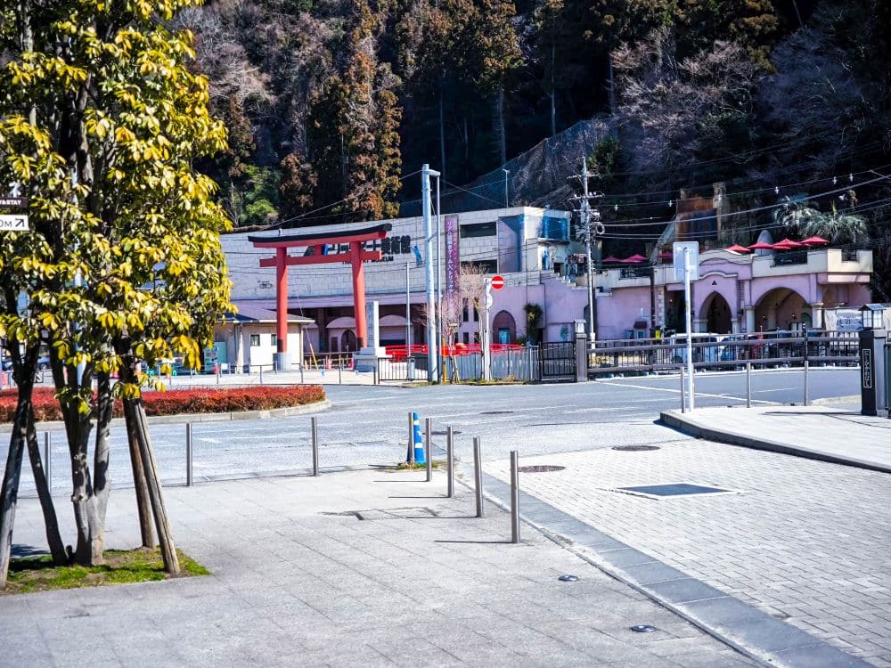 Eine ruhige Straße in der Nähe von Takao mit einem roten Torii-Tor, rosa Gebäuden und Bäumen im Hintergrund an einem sonnigen Tag in Japan. Metallgeländer säumen den Gehweg, und es sind nur wenige Menschen unterwegs.