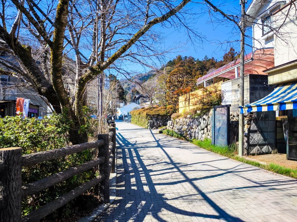 Eine ruhige, sonnenbeschienene Straße in Takao, einer kleinen Stadt mit Steinmauern, schattenwerfenden Bäumen, einem Holzzaun und Häusern auf beiden Seiten. Der Himmel ist klar und blau, und im Hintergrund sind Hügel zu sehen.