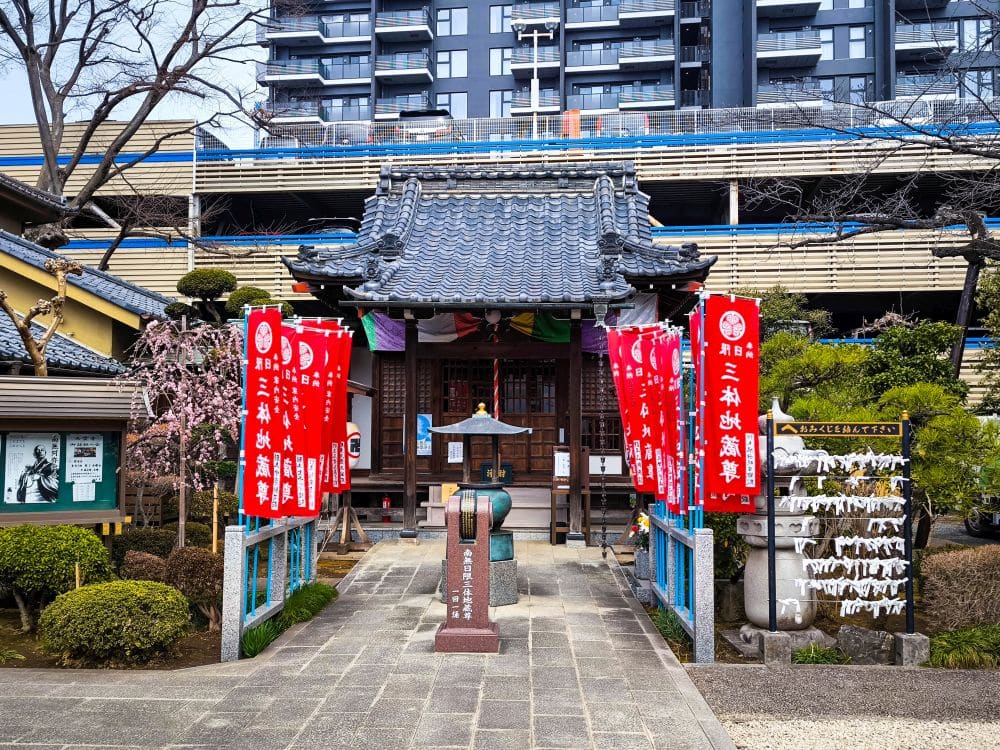 Ein kleiner japanischer Schrein mit einem traditionellen Ziegeldach in Kawagoe wird von roten Bannern und üppigem Grün flankiert und steht vor einem modernen Gebäude und einem Parkhaus. Weiße Papierglücksbringer hängen an einem Gestell auf der rechten Seite.