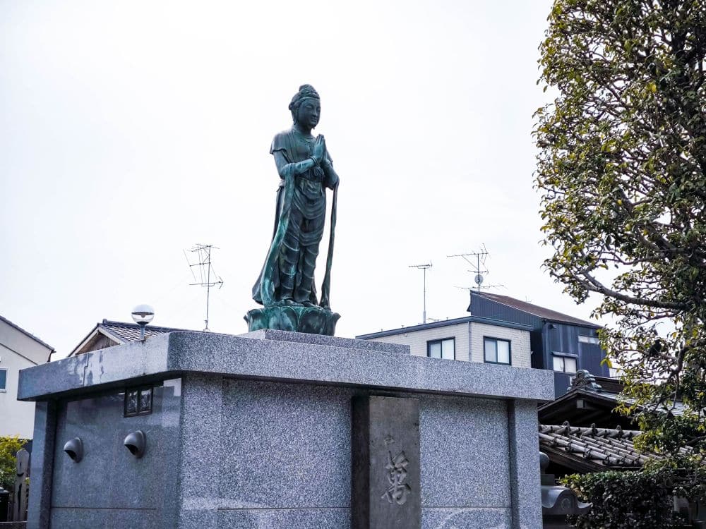 Eine Bronzestatue einer stehenden Figur in traditioneller Kleidung, die einen Stab hält, steht auf einem Steinsockel in Kawagoe. Im Hintergrund sind Wohnhäuser und Bäume zu sehen.