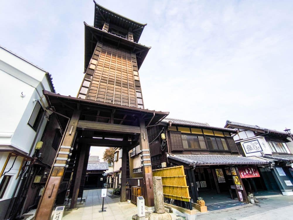 Ein hoher hölzerner Uhrenturm, bekannt als Toki no Kane, erhebt sich über traditionellen japanischen Gebäuden mit Ziegeldächern unter einem teilweise bewölkten Himmel im historischen Viertel von Kawagoe.