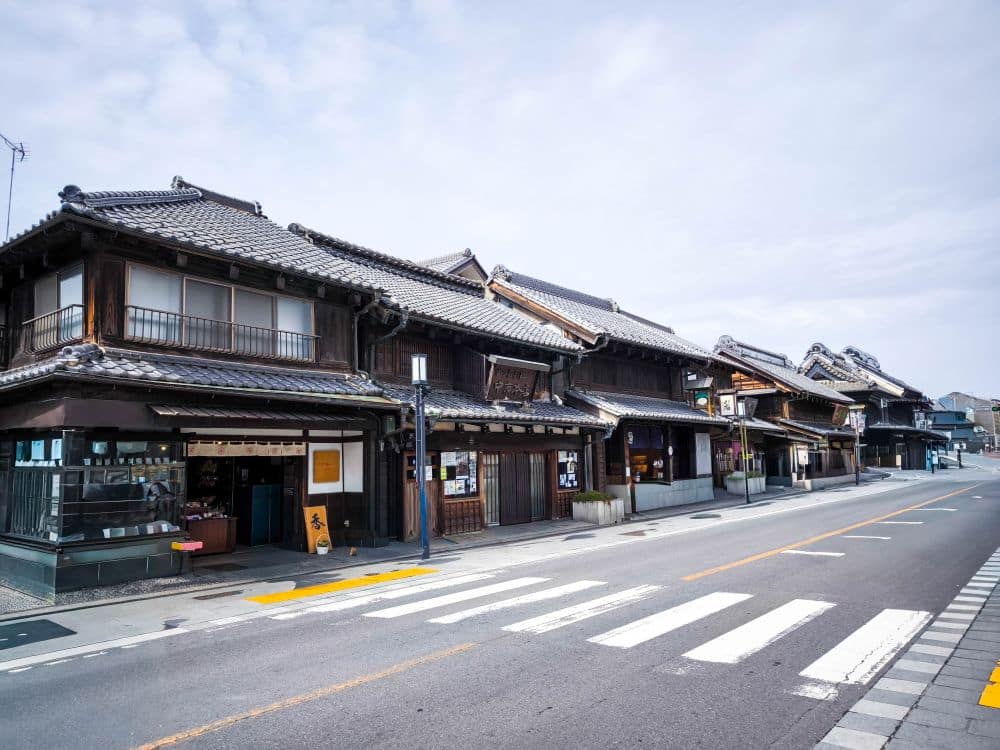 Eine Reihe traditioneller japanischer Holzhäuser säumt eine ruhige Straße mit einem Zebrastreifen unter einem bewölkten Himmel in Kawagoe. Die leere, gut gepflegte Straße fängt den friedlichen, historischen Charme dieser malerischen Stadt ein.