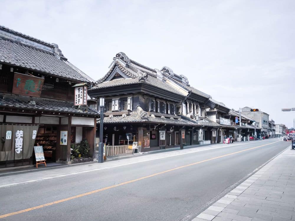Eine ruhige Straße in Kawagoe, gesäumt von traditionellen japanischen Gebäuden mit Ziegeldächern und Holzfassaden unter einem blassen, teilweise bewölkten Himmel. Die Gegend wirkt ruhig, ohne sichtbare Menschen oder Verkehr.