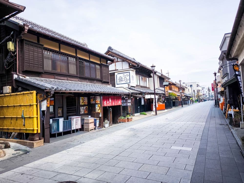 Eine ruhige, leere Straße in Kawagoe, gesäumt von traditionellen japanischen Holzhäusern und Ladenschildern mit Ziegeldächern und bunten Bannern unter einem hellen, klaren Himmel.