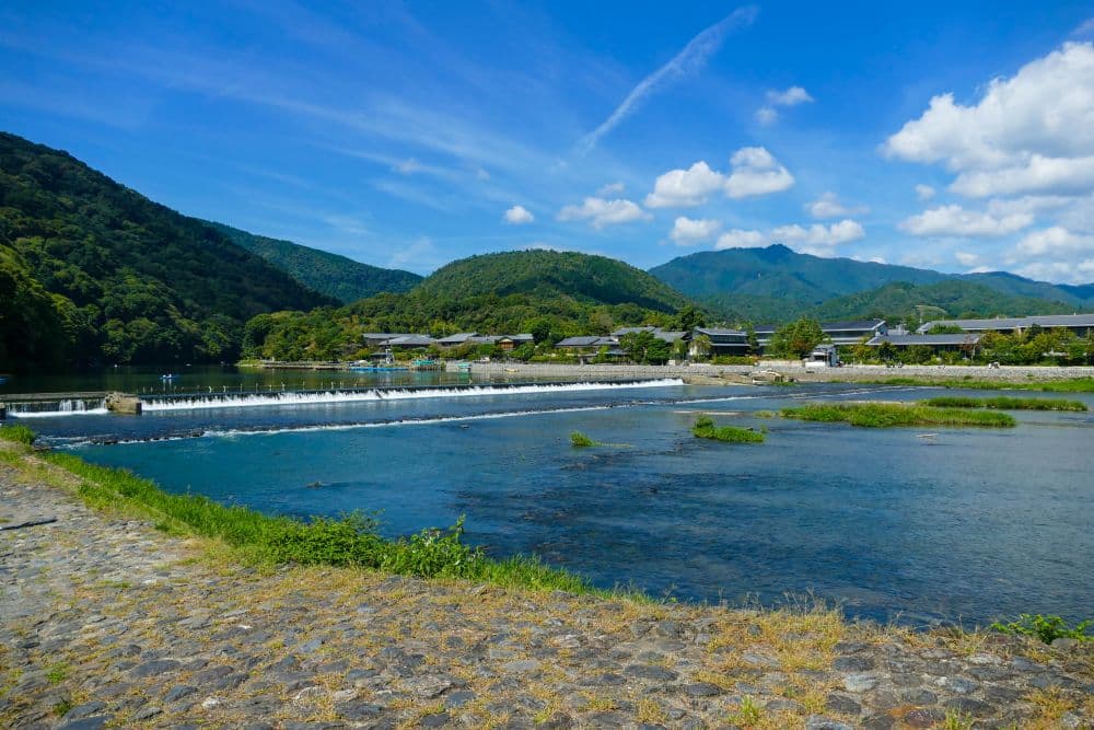 Ein malerischer Fluss fließt sanft über ein niedriges Wehr in Arashiyama, umgeben von üppig grünen Hügeln und traditionellen Gebäuden unter einem strahlend blauen Himmel mit vereinzelten Wolken. Ein Steinweg säumt das Flussufer im Vordergrund.