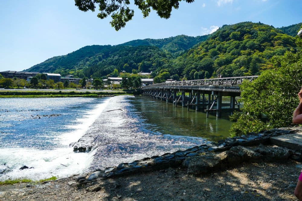 Eine Holzbrücke überspannt einen Fluss mit einem kleinen Wasserfall, umgeben von üppig grünen Hügeln und Bäumen unter einem klaren blauen Himmel, der an die landschaftliche Schönheit von Arashiyama erinnert. In der Ferne sind einige Gebäude zu sehen.