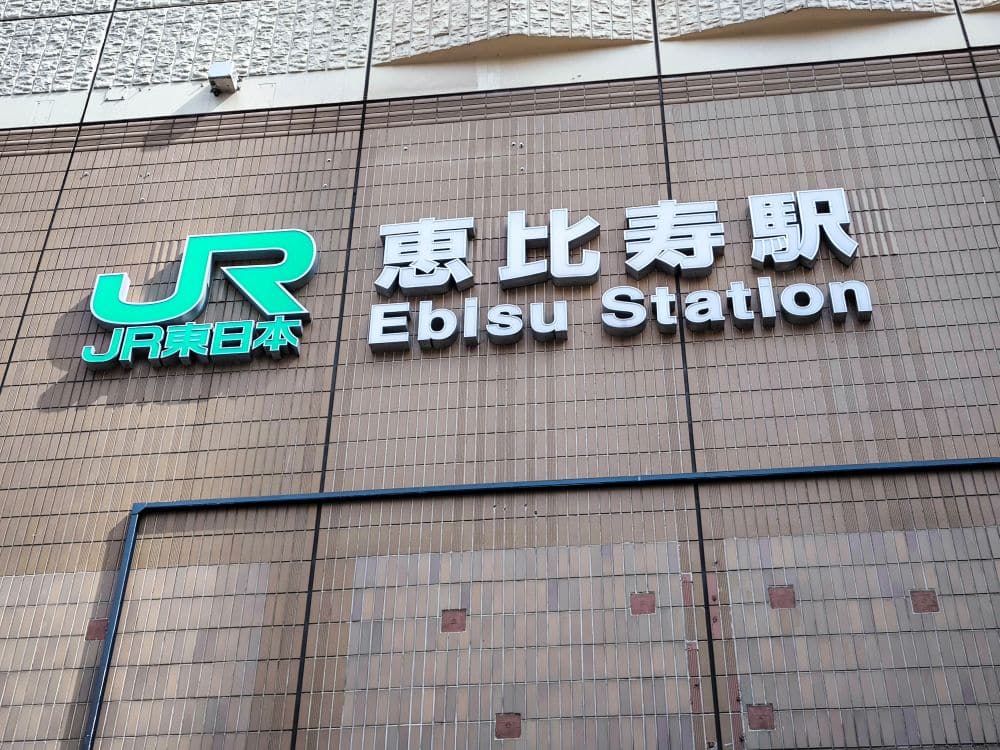 Eine Wand mit dem grün-weißen Logo von JR East Japan, daneben japanische Schriftzeichen und der englische Schriftzug „Ebisu Station“. Die beige geflieste Fassade spiegelt die Verbindung zur geschäftigen Yamanote-Linie wider.