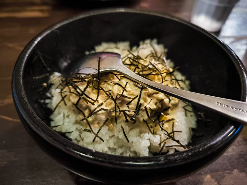 Eine schwarze Hino-Schale, gefüllt mit weißem Reis, bedeckt mit einer cremigen Sauce und dünnen Algenstreifen, auf der ein Metalllöffel ruht. Die Schale steht auf einem Holztisch.