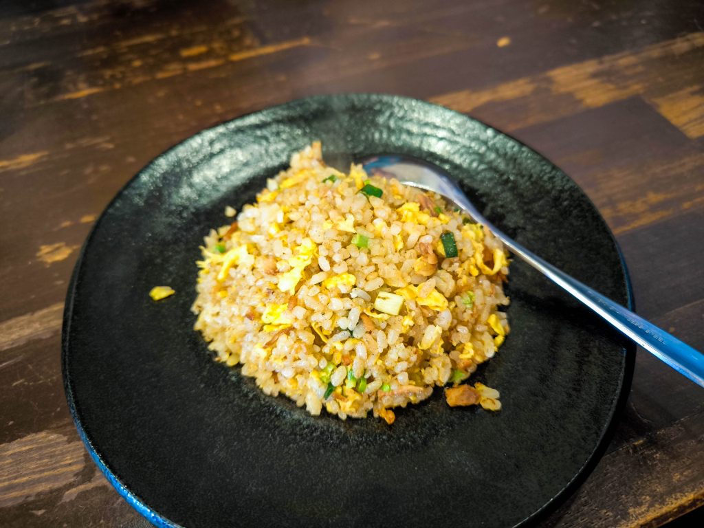 Ein schwarzer Teller mit einer Portion gebratenem Reis aus Hino, gemischt mit gehackten grünen Zwiebeln und Rührei. Ein Metalllöffel liegt auf dem Rand des Tellers vor dem Hintergrund eines Holztisches.