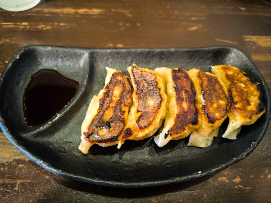 Fünf in der Pfanne gebratene Teigtaschen, eine Spezialität aus Hino, liegen in einer Reihe auf einem schwarzen Teller mit einer kleinen Portion Sojasauce an der Seite auf einem dunklen Holztisch.