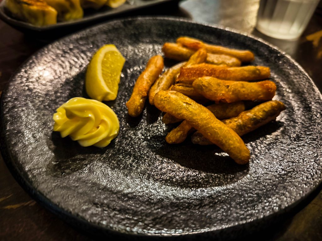 Ein schwarzer Teller mit gebratenen kleinen Fischen, einer Zitronenspalte und einem Strudel Mayonnaise steht auf einem dunklen Tisch im Hino. Im Hintergrund stehen ein verschwommener Teller und ein Glas Wasser.