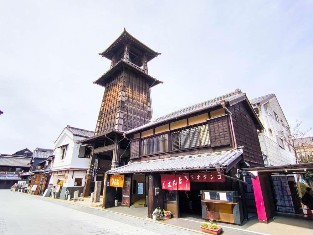Der hohe hölzerne Uhrenturm Toki no Kane erhebt sich über den traditionellen japanischen Gebäuden mit Ziegeldächern in Kawagoe. Geschäfte mit roten Schildern säumen die Straße darunter und schaffen an diesem sonnigen Tag eine historische Stadtatmosphäre.