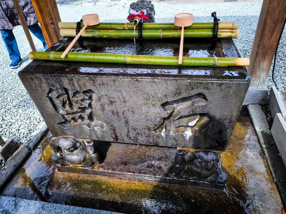 Ein japanischer Reinigungsbrunnen in Kawagoe mit Bambuskellen, die auf einem Steinbecken ruhen, mit geschnitzten Kanji-Zeichen und dekorativen geschnitzten Figuren am Sockel.