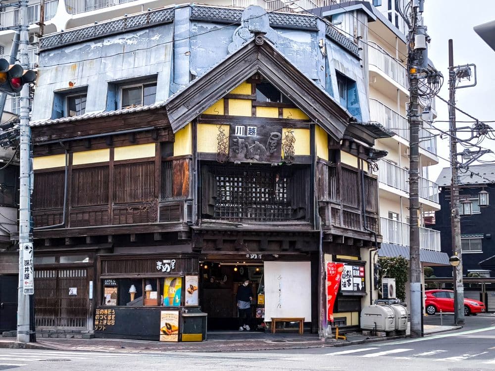 Ein traditionelles japanisches Holzgebäude mit einem Laden im Erdgeschoss steht an einer Straßenecke in Kawagoe, umgeben von modernen Gebäuden und Stromleitungen.