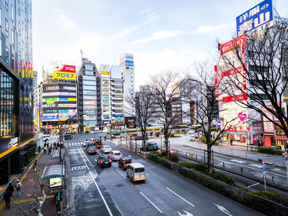 Eine belebte Stadtstraße in Japan mit vielen Autos und Bussen kreuzt die geschäftige Yamanote-Linie. Hohe Gebäude, geschmückt mit bunten Werbetafeln und Schildern, säumen beide Seiten. Kahle Bäume lassen eine Winterlandschaft unter einem klaren, von vereinzelten Wolken gesprenkelten Himmel erahnen.