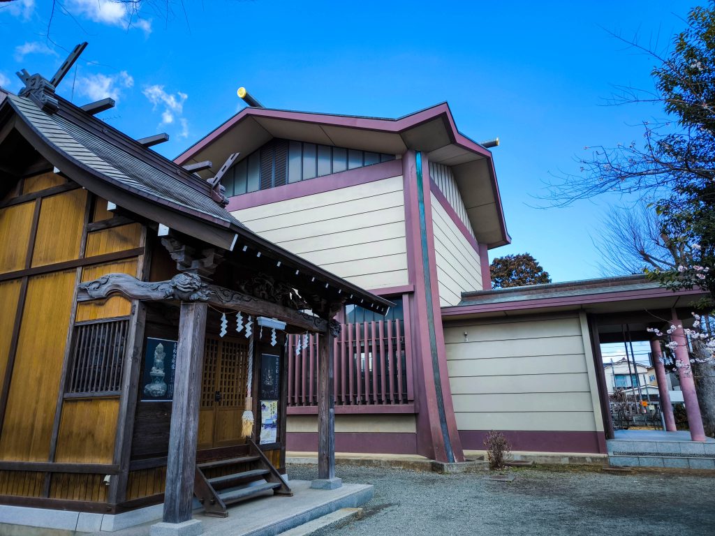 Ein traditioneller japanischer Schrein mit Holzstrukturen steht neben einem modernen Gebäude in Hino, beide unter einem klaren blauen Himmel. Im Vordergrund sind einige Bäume und eine Kiesfläche zu sehen, die die Geschichte mit dem Geist des modernen Tokio verbinden.
