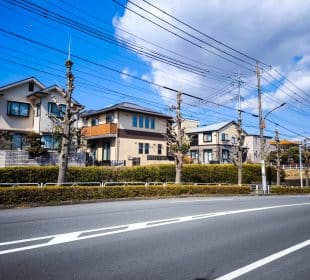 Eine ruhige Vorstadtstraße in Hino, Japan, mit ordentlich beschnittenen Bäumen, modernen Häusern, Stromleitungen über dem Haus und einem klaren blauen Himmel. Hecken trennen die Häuser von der Straße und spiegeln eine friedliche Harmonie wider, die an einen alten Samurai-Tempel erinnert.