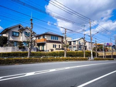 Eine ruhige Vorstadtstraße in Hino, Japan, mit ordentlich beschnittenen Bäumen, modernen Häusern, Stromleitungen über dem Haus und einem klaren blauen Himmel. Hecken trennen die Häuser von der Straße und spiegeln eine friedliche Harmonie wider, die an einen alten Samurai-Tempel erinnert.