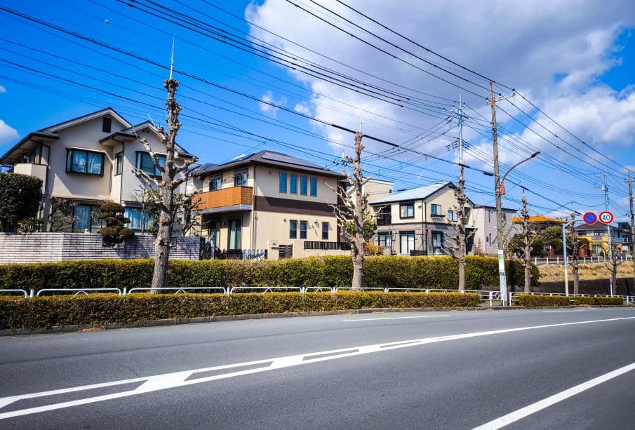 Eine ruhige Vorstadtstraße in Hino, Japan, mit ordentlich beschnittenen Bäumen, modernen Häusern, Stromleitungen über dem Haus und einem klaren blauen Himmel. Hecken trennen die Häuser von der Straße und spiegeln eine friedliche Harmonie wider, die an einen alten Samurai-Tempel erinnert.