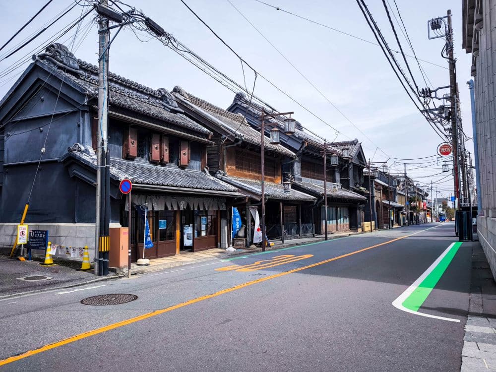 Eine ruhige Straße in Kawagoe, Japan, gesäumt von traditionellen Holzhäusern, Überlandleitungen und Straßenmarkierungen. Die Gegend wirkt leer und friedlich unter einem hellen, klaren Himmel.
