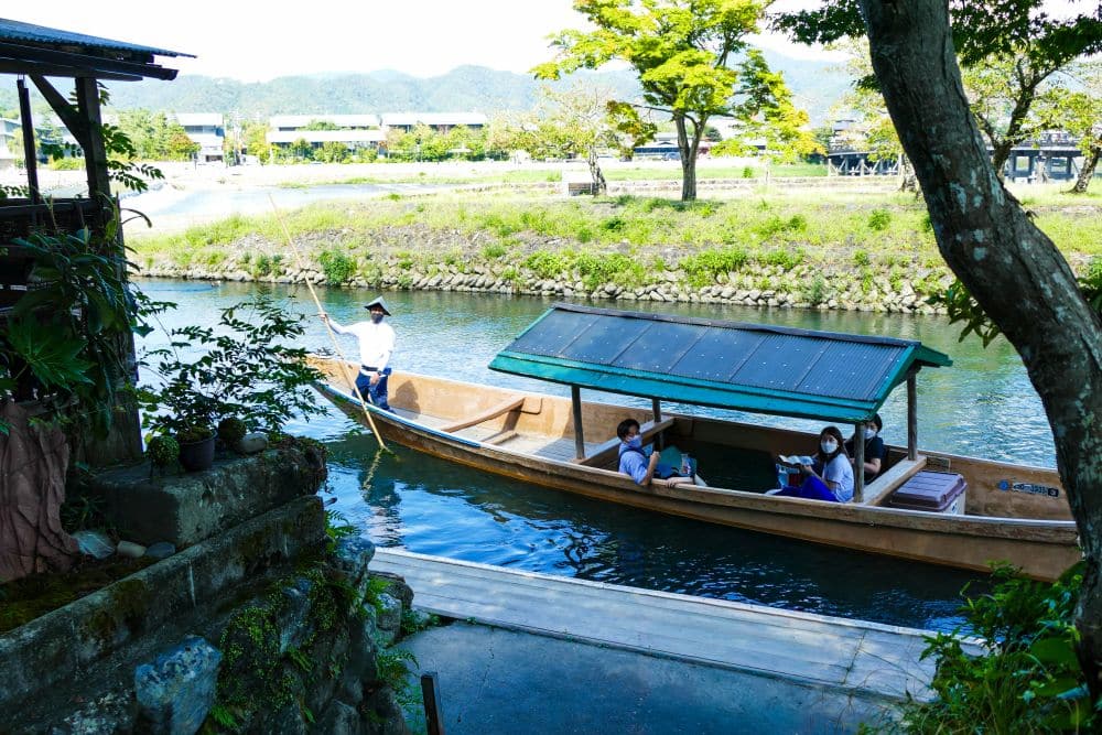 Ein kleines Holzboot mit einem grünen Dach gleitet auf einem ruhigen Fluss in Arashiyama. Eine Person in traditioneller Kleidung steuert es, während maskierte Passagiere darin sitzen. Bäume und üppiges Grün säumen das Flussufer unter einem klaren Himmel.