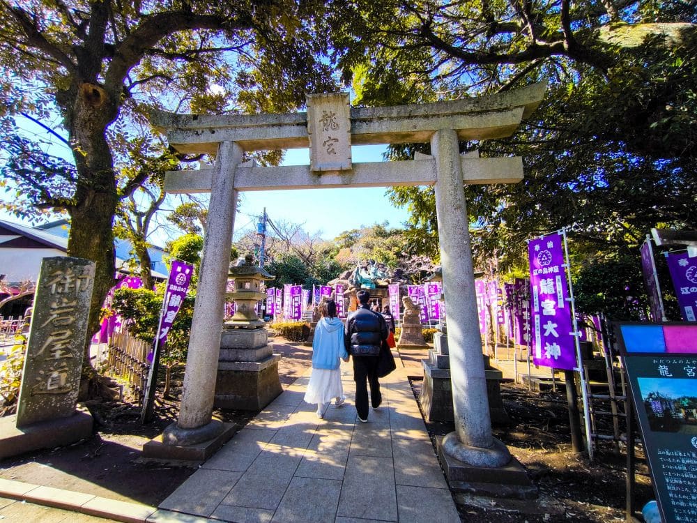 Zwei Personen gehen an einem sonnigen Tag unter dem traditionellen Torii-Tor in Okutsunomiya, einem japanischen Schrein, der von Bäumen und violetten Bannern umgeben ist.