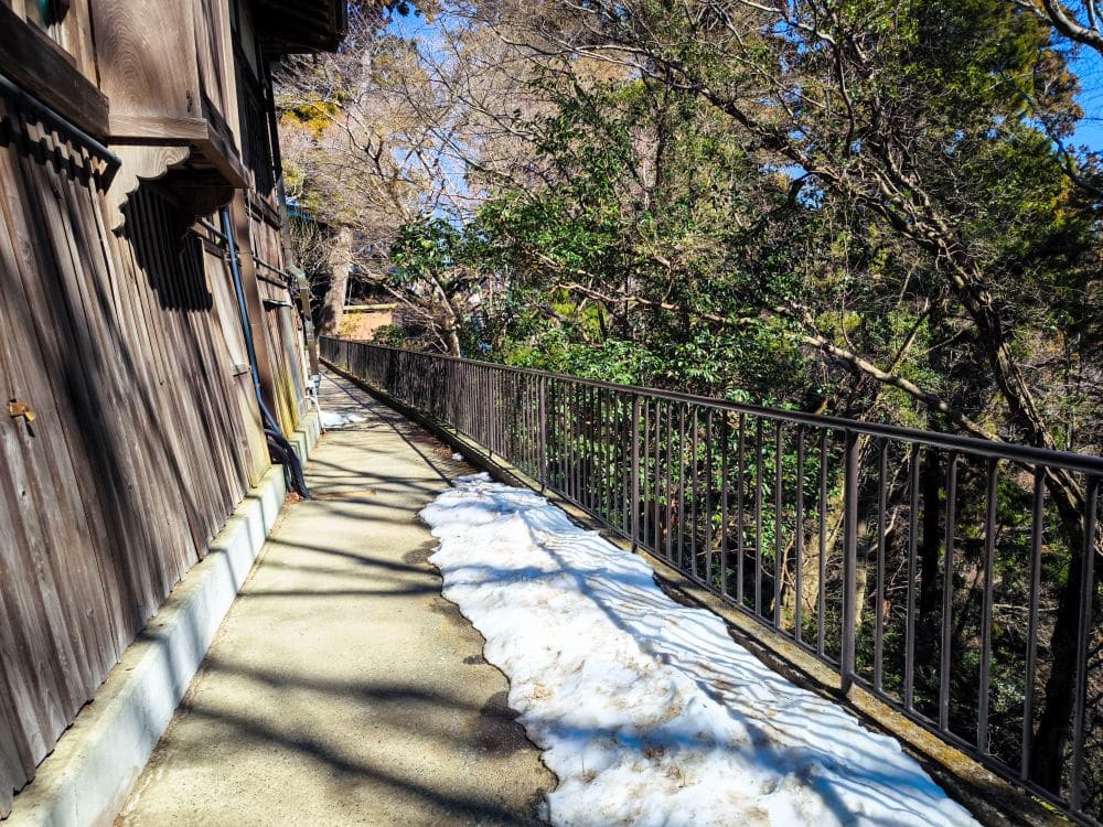 Ein schmaler Betonweg in Takao führt an einem Holzgebäude mit einem Metallgeländer entlang, das von Bäumen gesäumt wird. Im hellen Sonnenlicht schmelzen die Schneeflecken auf dem Gehweg.