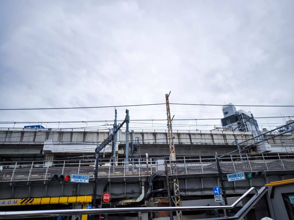 Die Hochbahngleise der Yamanote-Linie erstrecken sich durch die Stadt, darunter sind Ampeln und Schilder verstreut. Ein bewölkter Himmel dient als Kulisse, während verschiedene Gebäude und Bauwerke das geschäftige Stadtbild prägen.