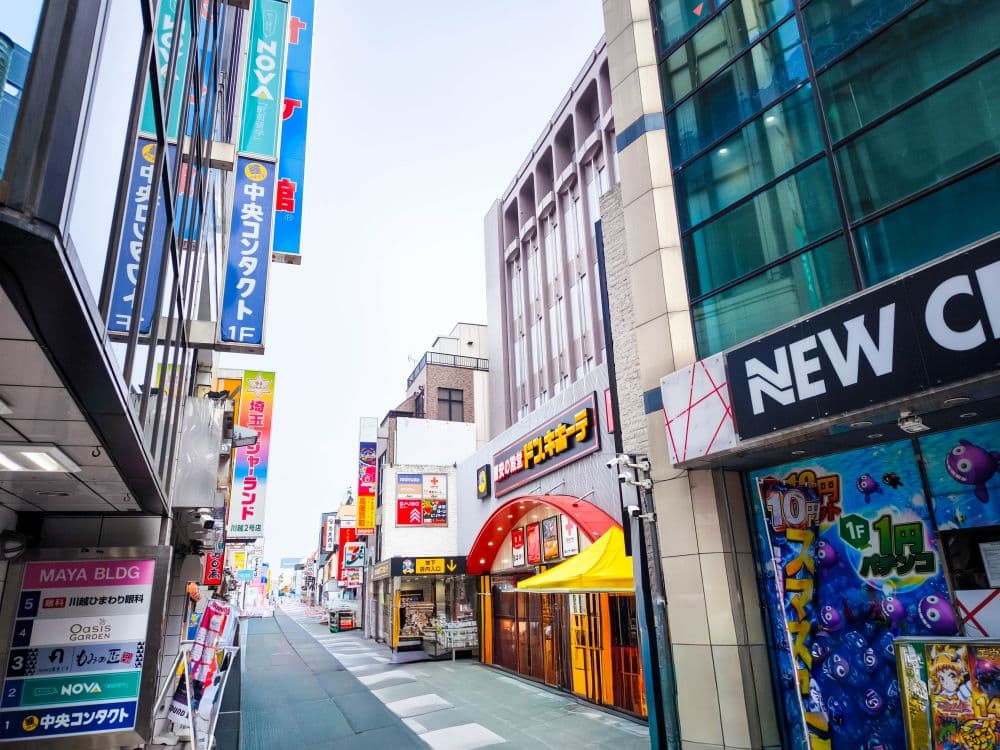Eine farbenfrohe Straße in Kawagoe, Japan, gesäumt von Geschäften und Schildern in japanischer Sprache, darunter eine leuchtend gelbe und rote Schaufensterfront und ein großes Schild mit der Aufschrift "NEW". Die Straße scheint frei von Fußgängern zu sein.