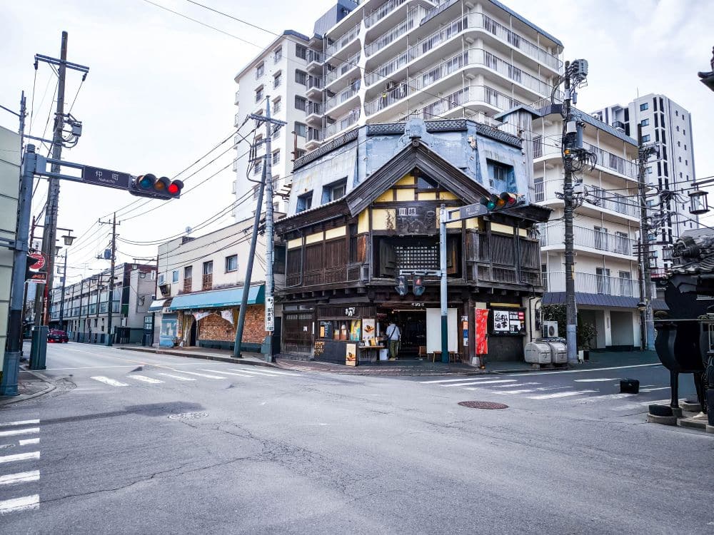 In einer ruhigen Straße in Kawagoe, Japan, steht ein traditionelles Holzgebäude an der Ecke, umgeben von modernen Wohnkomplexen. Über dem Gebäude hängen Ampeln und die Straße ist leer von Autos und Menschen.