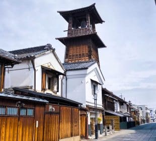 Eine traditionelle japanische Straße in Kawagoe mit Holzhäusern und einem hohen, historischen Uhrenturm aus Holz unter einem bewölkten Himmel. Die ruhige, leere Straße erweckt ein Gefühl von Ruhe und Nostalgie.