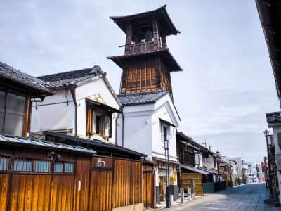Eine traditionelle japanische Straße in Kawagoe mit Holzhäusern und einem hohen, historischen Uhrenturm aus Holz unter einem bewölkten Himmel. Die ruhige, leere Straße erweckt ein Gefühl von Ruhe und Nostalgie.
