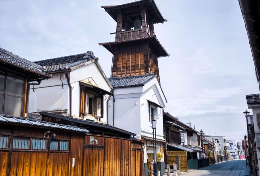 Eine traditionelle japanische Straße in Kawagoe mit Holzhäusern und einem hohen, historischen Uhrenturm aus Holz unter einem bewölkten Himmel. Die ruhige, leere Straße erweckt ein Gefühl von Ruhe und Nostalgie.