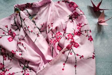 Ein rosafarbener Kimono mit Kirschblütenmuster ist auf einer hellen Fläche ausgebreitet. Zwei rosafarbene Origamipapierkraniche liegen daneben und ergänzen die Eleganz des Kimonos perfekt.