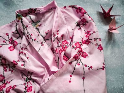 Ein rosafarbener Kimono mit Kirschblütenmuster ist auf einer hellen Fläche ausgebreitet. Zwei rosafarbene Origamipapierkraniche liegen daneben und ergänzen die Eleganz des Kimonos perfekt.