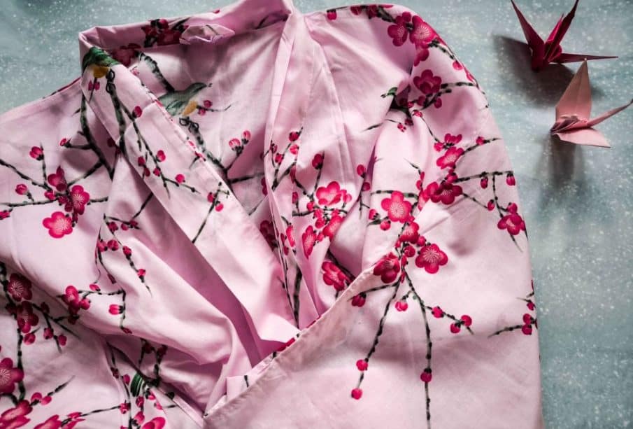 Ein rosafarbener Kimono mit Kirschblütenmuster ist auf einer hellen Fläche ausgebreitet. Zwei rosafarbene Origamipapierkraniche liegen daneben und ergänzen die Eleganz des Kimonos perfekt.