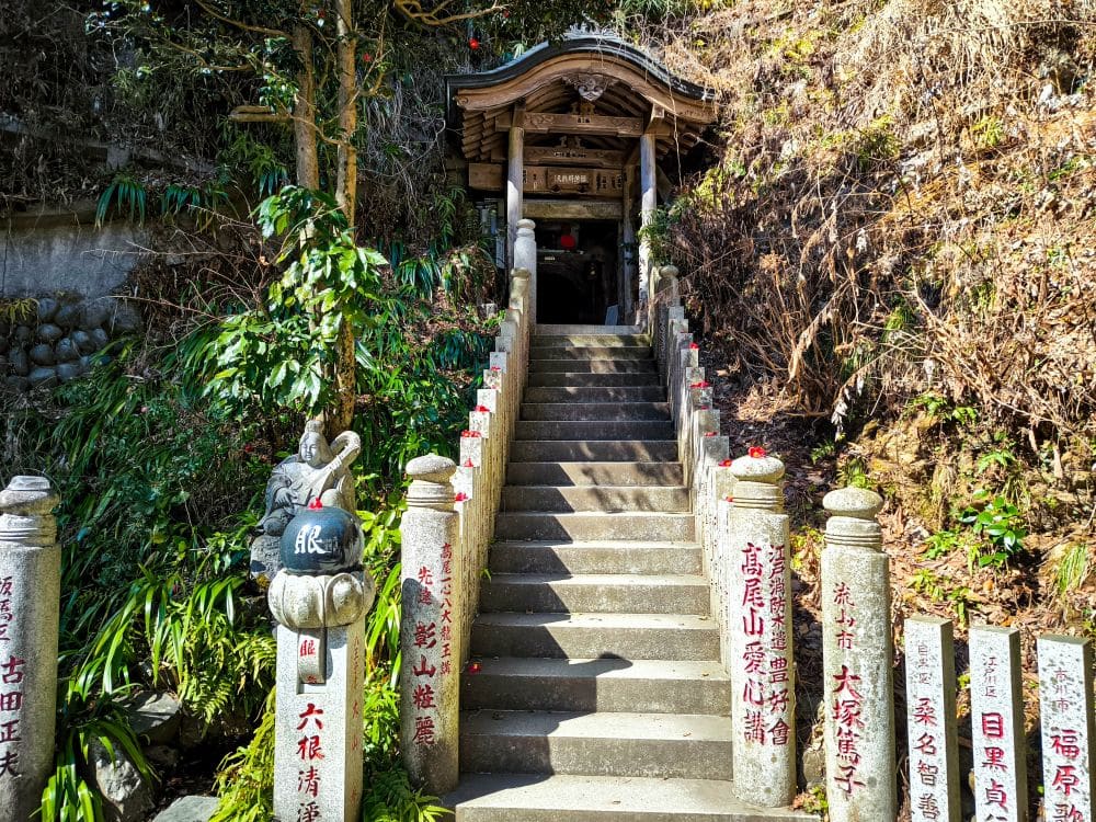 Eine Steintreppe mit rotem Geländer führt hinauf zu einem kleinen Holzschrein auf Takao, der von üppigem Grün und Steinsäulen mit eingravierten japanischen Schriftzeichen umgeben ist. Das Sonnenlicht erhellt die Stufen und die Vegetation.