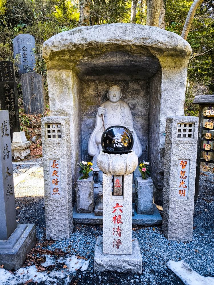 Eine steinerne Statue einer buddhistischen Figur befindet sich in einem kleinen Schrein auf Takao, umgeben von geschnitzten Säulen mit japanischen Inschriften. Eine verzierte schwarze Kugel ruht auf einem Sockel davor, und Blumen sind als Opfergaben aufgestellt.