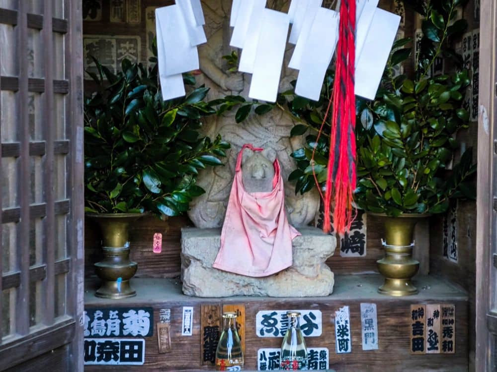 In einem hölzernen Schrein auf Takao, der mit japanischen Postern und Flaschen geschmückt ist, sitzt eine kleine Steinstatue mit einem verblichenen rosa Lätzchen zwischen zwei goldenen Vasen mit grünen Zweigen, umgeben von Papiertalismanen und bunten Seilen.