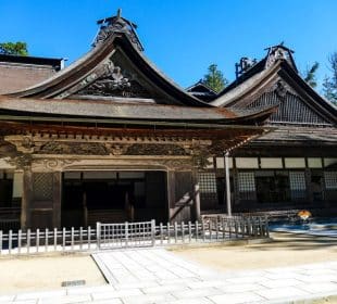 Traditioneller japanischer Holztempel mit komplizierten Dachformen und Schnitzereien vor einem strahlend blauen Himmel. Dies ist der Kongobu-ji Tempel in Koyasan, der oft als das spirituelle Herz Japans bezeichnet wird, mit Bäumen im Hintergrund und einem kleinen Holzzaun am Eingang.
