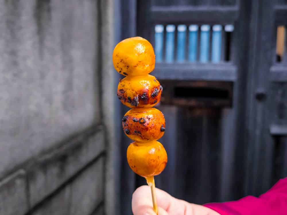 Eine Hand hält einen Spieß mit vier gegrillten, leicht verkohlten Dangokugeln vor einem unscharfen, rustikalen Hintergrund aus Holztür und Betonwand in Kawagoe.