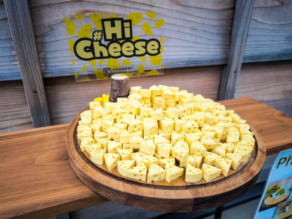 Auf einer runden Holzplatte liegen viele kleine, gelbe, keilförmige Käsestücke. Hinter der Platte steht ein Schild mit der Aufschrift #Hi Cheese vor einem Hintergrund aus Käse-Illustrationen, was der Auslage eine charmante Kawagoe-Note verleiht.