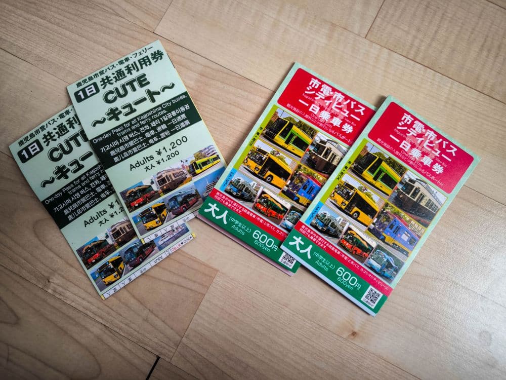 Vier Kagoshima-Bus-Tageskarten mit Fotos von Bussen und Stadtansichten sind auf einem Holzboden ausgestellt. Auf den Fahrkarten sind die Preise für Erwachsene (1.200 Yen und 600 Yen) angegeben und der Text ist auf Japanisch und etwas Englisch.