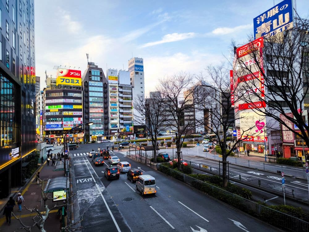 Eine belebte Straße in der Nähe von Haneda in Japan mit Autos, Fußgängern, blattlosen Bäumen und hohen Gebäuden mit bunten Schildern und Werbetafeln unter einem teilweise bewölkten Himmel.