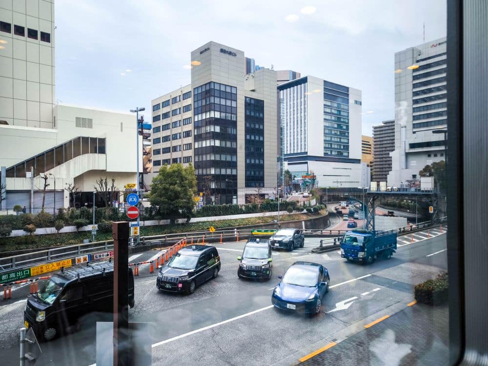 Eine belebte Straße in der Nähe von Haneda mit Autos, Taxis und einem blauen Lastwagen, die an modernen Bürogebäuden unter bewölktem Himmel vorbeifahren. Auf dem Fenster, durch das das Foto aufgenommen wurde, sind Reflexionen der Innenbeleuchtung zu sehen.