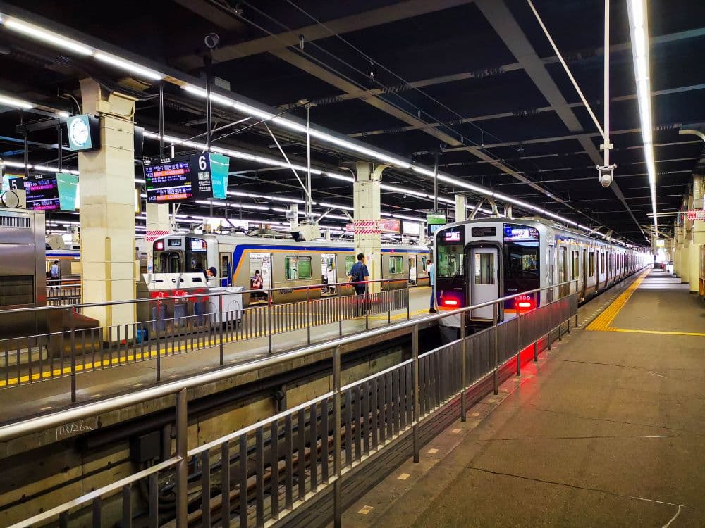 Ein hell erleuchteter Bahnsteig des japanischen Bahnhofs Haneda mit zwei angehaltenen Zügen, einsteigenden Fahrgästen und Informationsschildern über dem Kopf. Der Bahnsteig ist sauber mit gelbem taktilen Pflaster entlang der Kante.