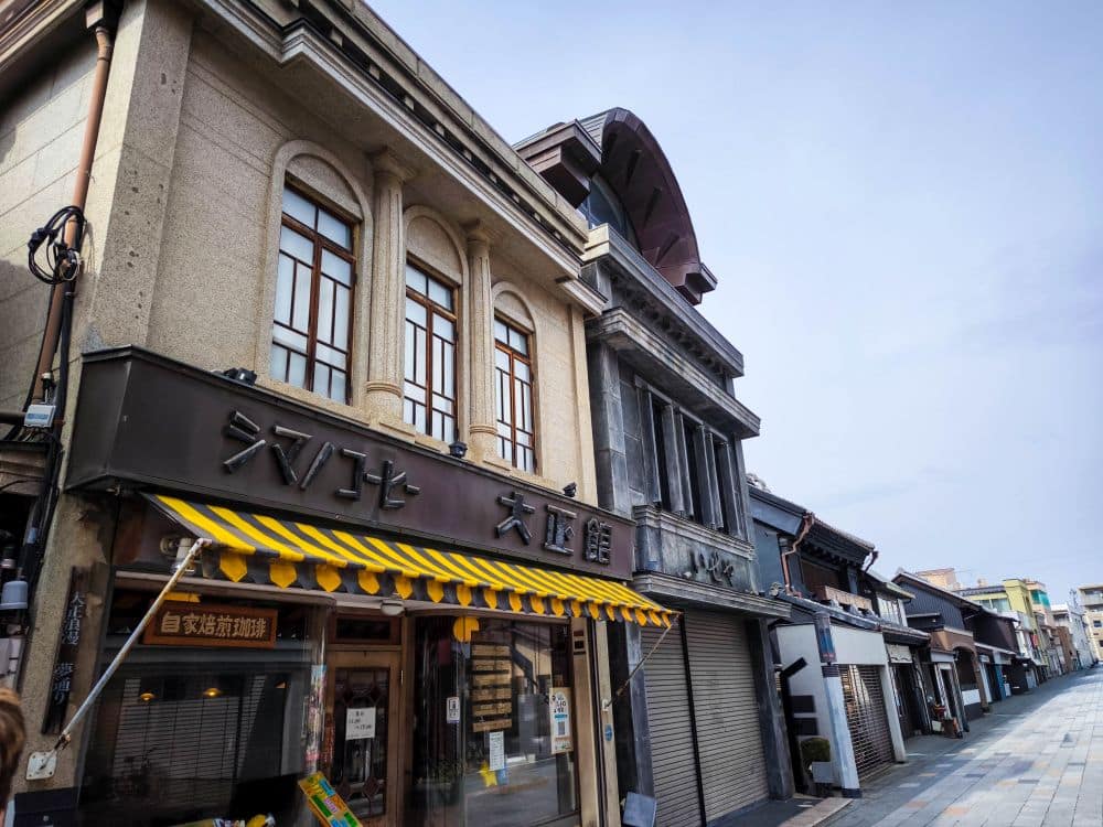 Eine ruhige Straße in Kawagoe mit traditionellen japanischen Gebäuden, eines davon mit einer gelb gestreiften Markise und großen Fenstern. Die Schilder sind auf Japanisch, einige Ladengeschäfte sind geschlossen, und der Himmel ist klar und blau.