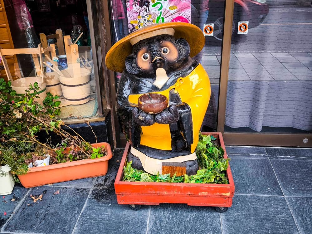Eine große Marderhund-Statue mit Strohhut und gelber Jacke hält eine Schüssel, während sie in einer roten Kiste mit Grünzeug steht, vor einem Ladeneingang auf dem gekachelten Bürgersteig von Kawagoe.
