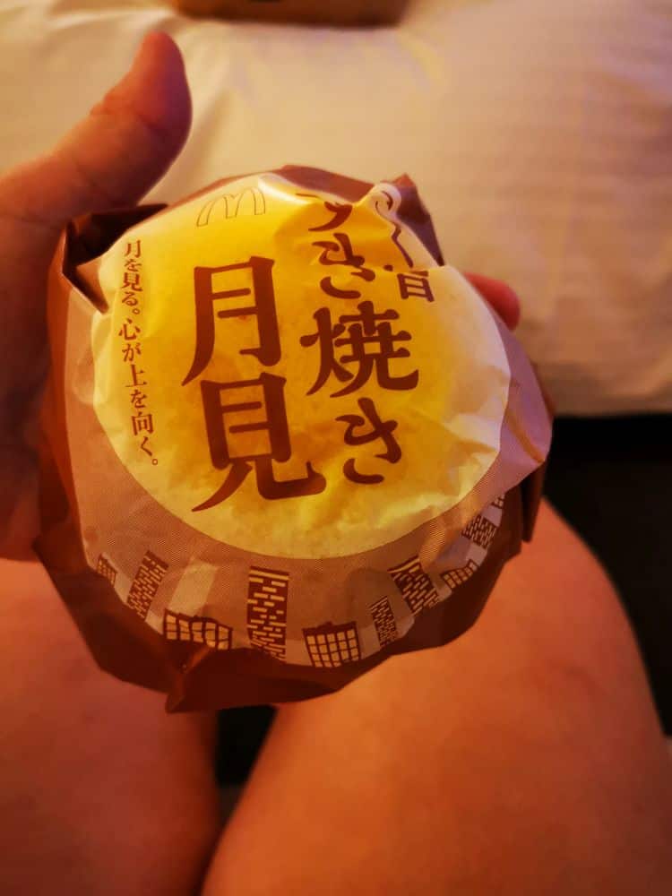 Eine Hand hält einen eingewickelten Burger mit japanischem Text auf der Verpackung, die einen gelben mondähnlichen Kreis und braune Akzente aufweist. Im Hintergrund ist ein Bett oder eine weiche Oberfläche zu sehen.