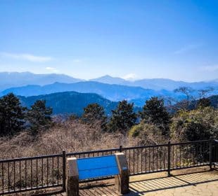 Von einem eingezäunten Aussichtspunkt mit einem Schild hat man einen herrlichen Blick auf die blauen Berge und Bäume - in der Ferne erhebt sich der schneebedeckte Gipfel von Takao unter einem klaren blauen Himmel.
